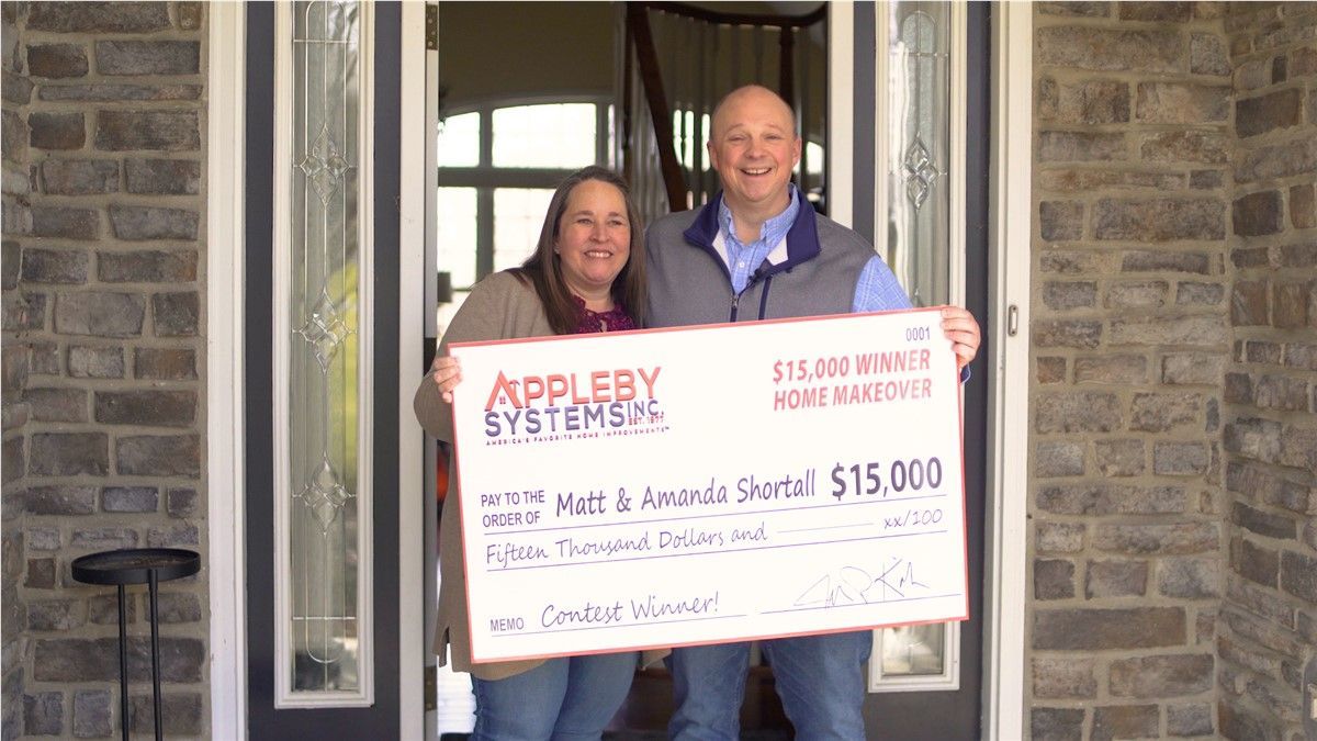 Two people hold a large $15,000 check at a front doorway, smiling for a photo.