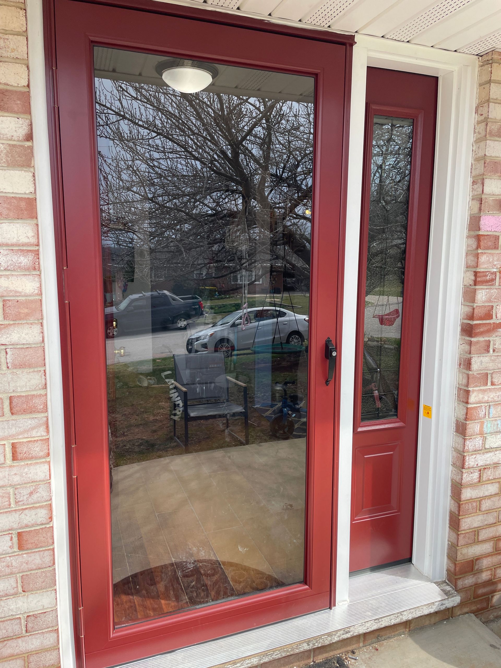 Red-framed glass front door with a narrow side window, reflecting trees and a chair outside.