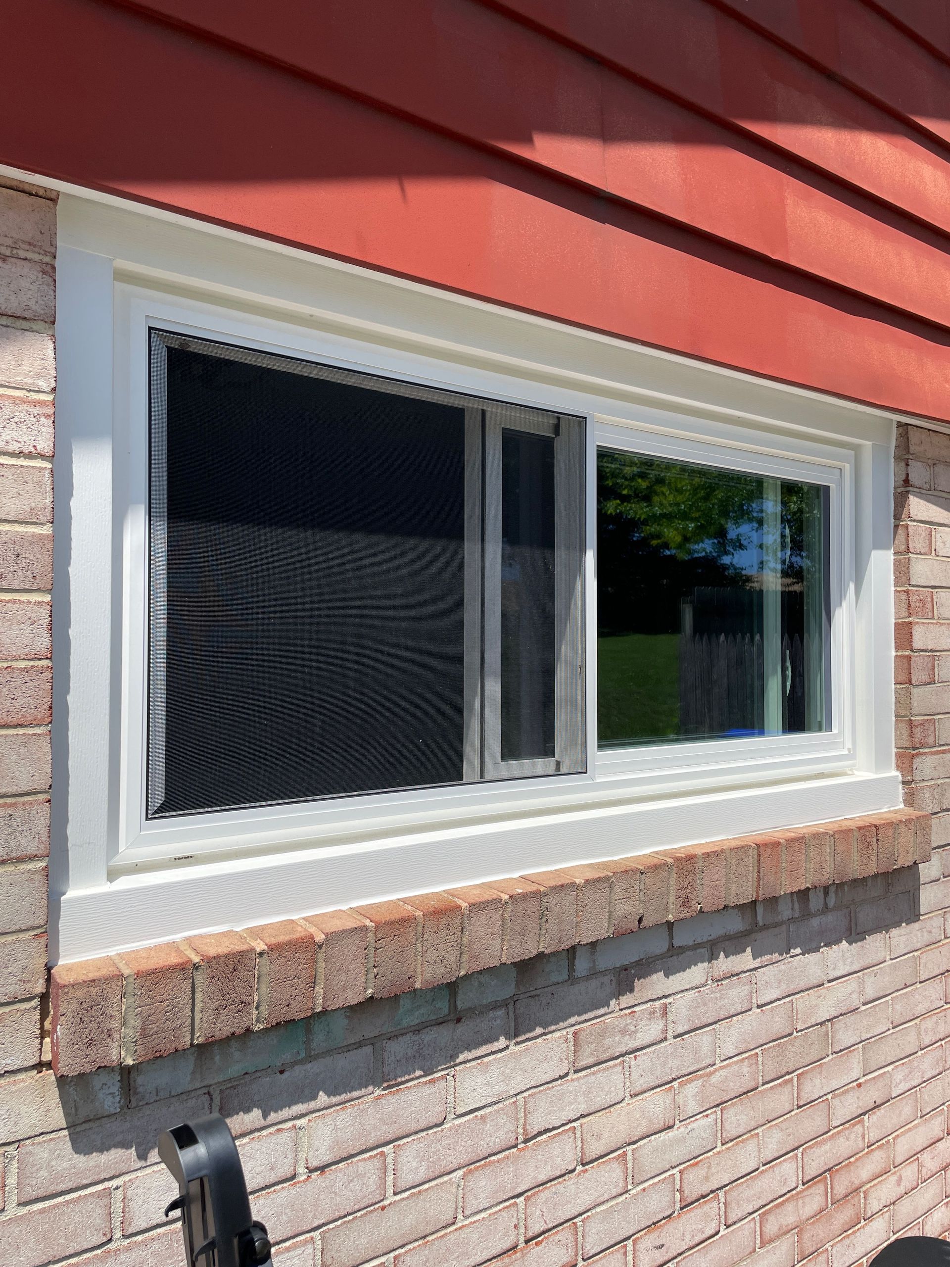White-framed sliding window with a screen, set in a brick wall beneath red siding.