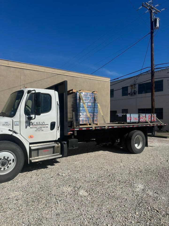 A white flatbed truck parked next to a building, loaded with pallets of bottled water and building materials under a clear blue sky.