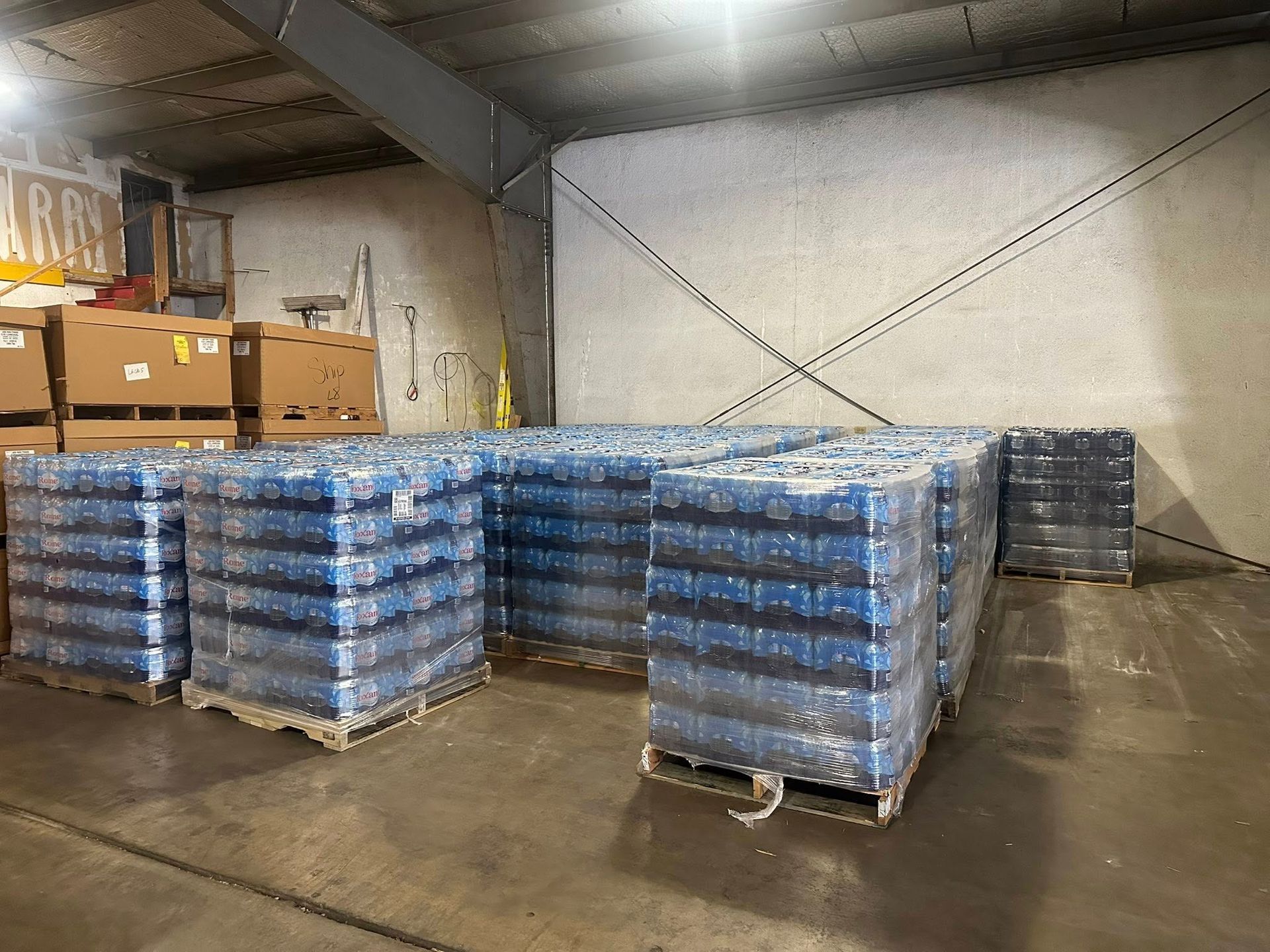 Pallets of bottled water stacked inside a warehouse, against a gray wall.