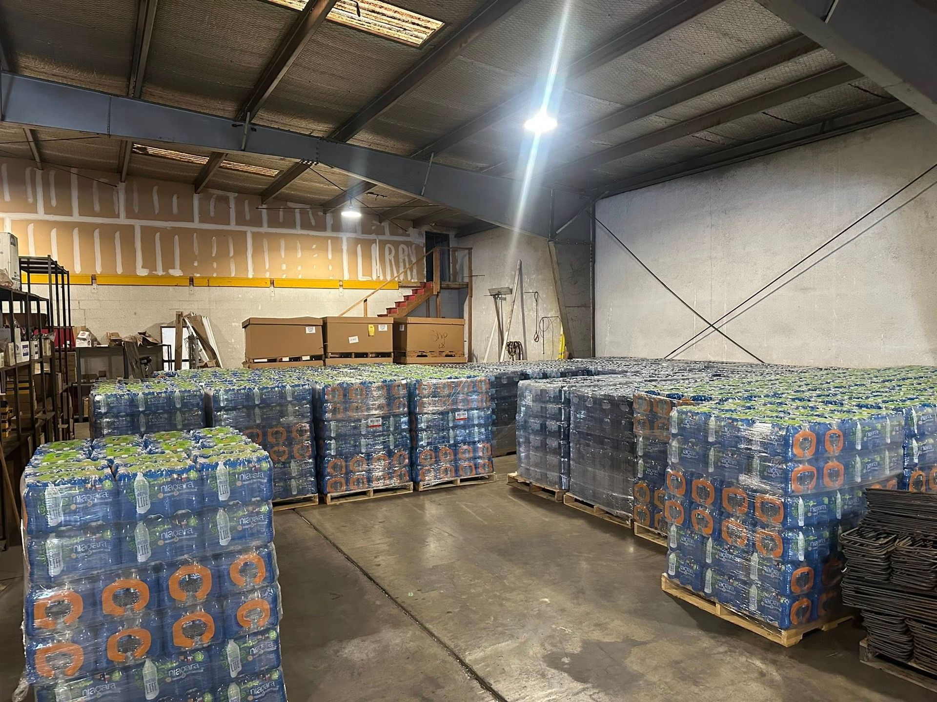 Warehouse interior with pallets of bottled water stacked. Light illuminates the corrugated ceiling.