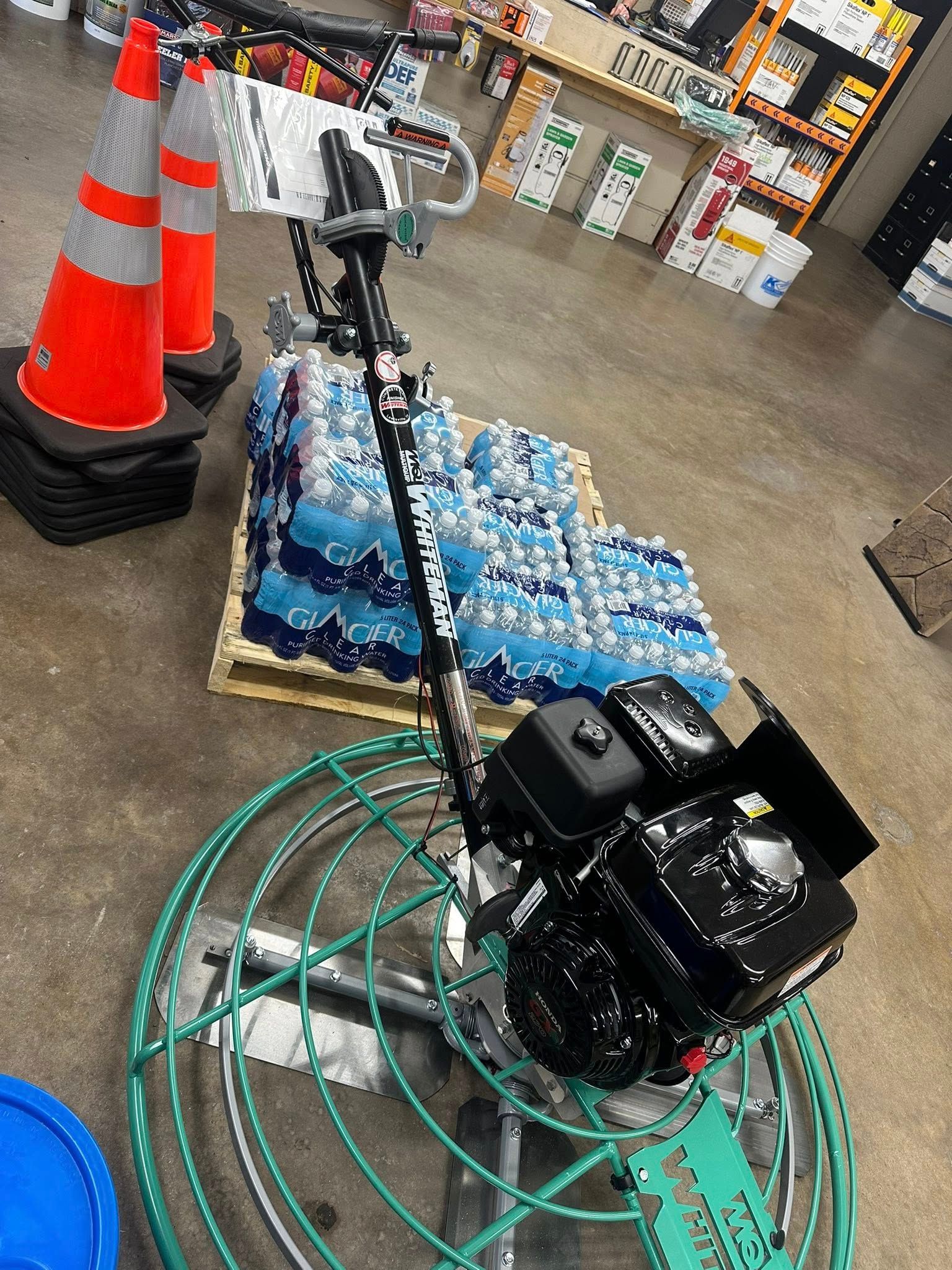 Concrete power trowel, black engine, atop a pallet of bottled water, orange traffic cones in background.