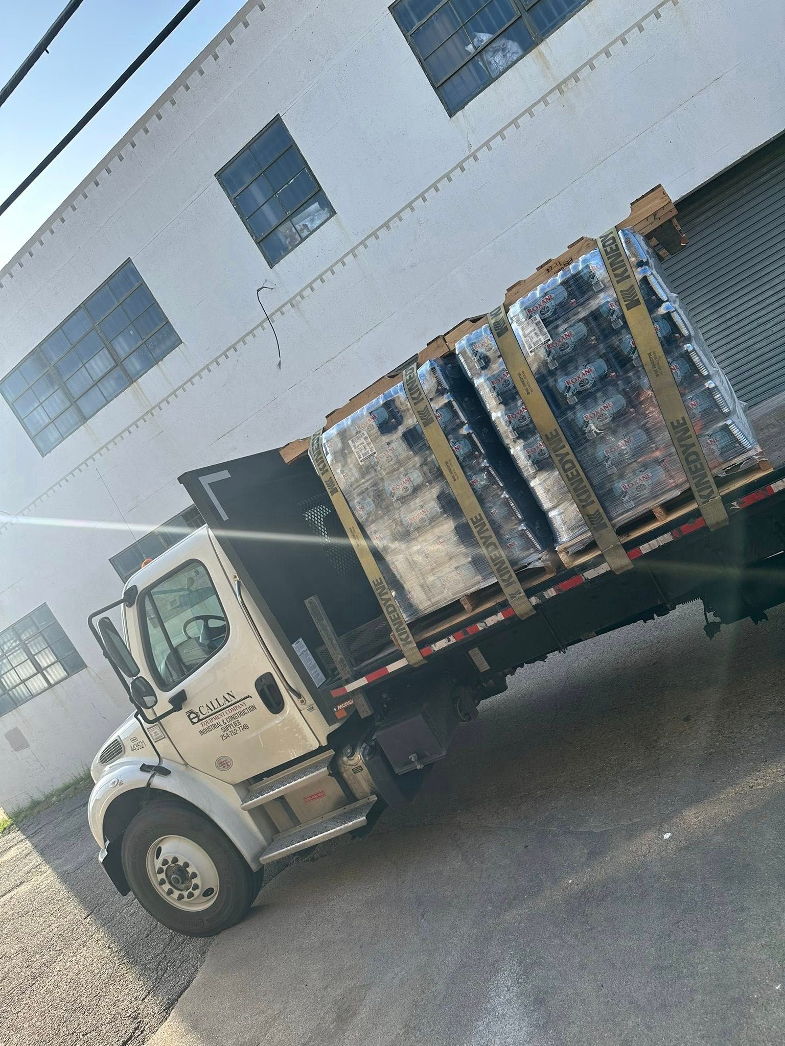 A flatbed truck carrying several wrapped pallets parked near a white building with open bay doors.