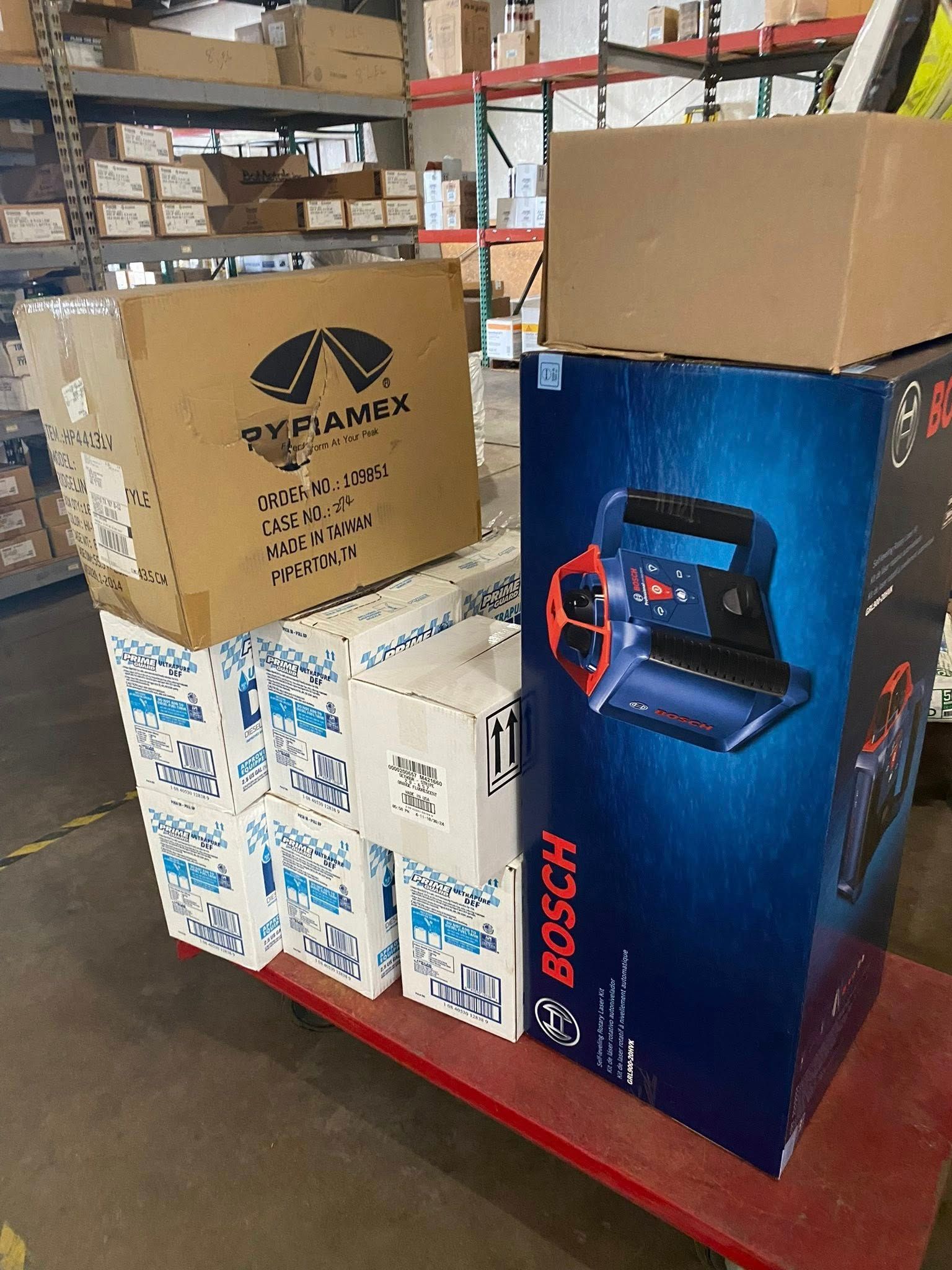 Boxes stacked on a red cart in a warehouse. Includes a Bosch power tool box and other boxes with logos.