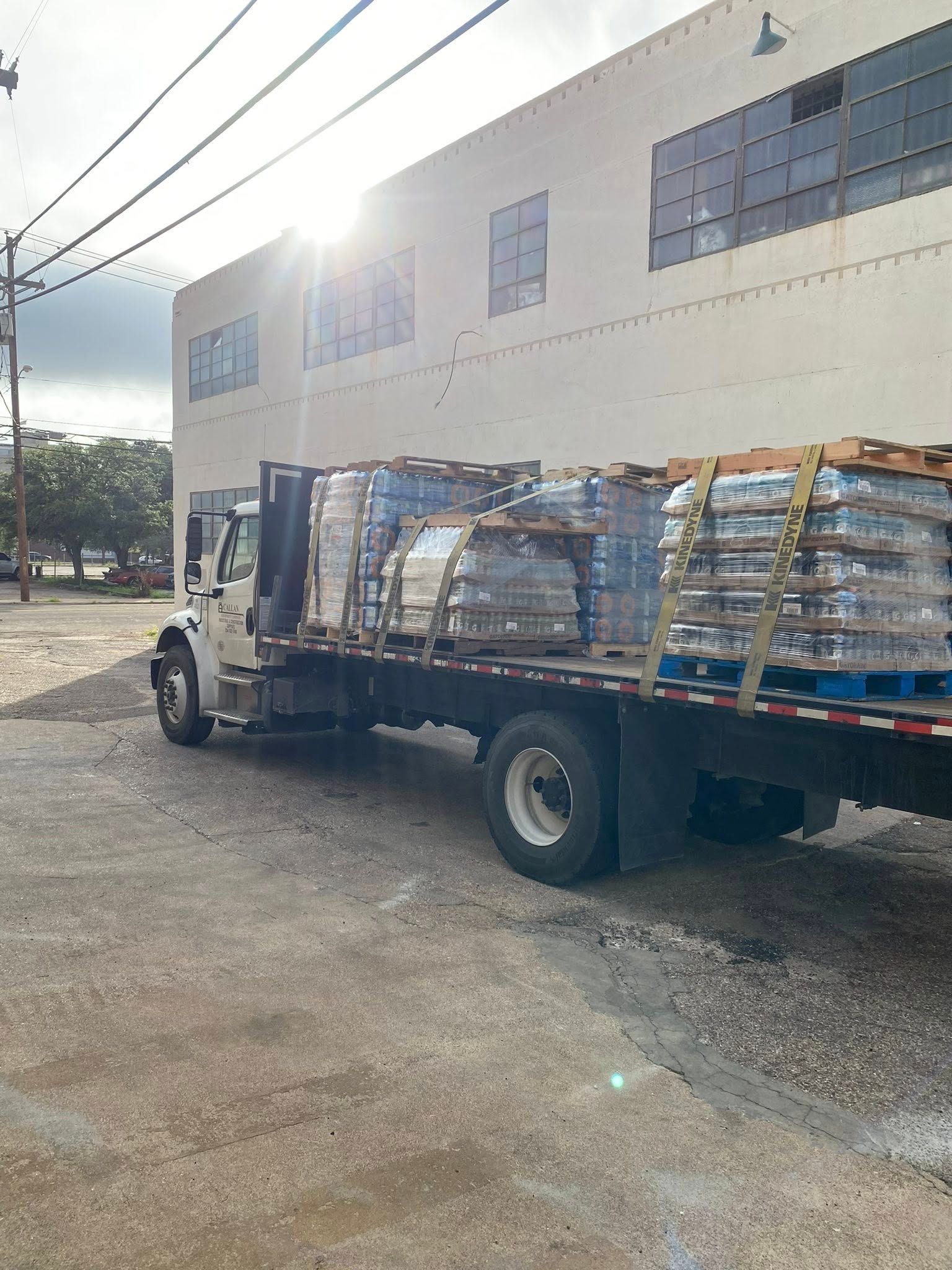 Truck loaded with pallets of bottled water in front of a white building on a bright day.