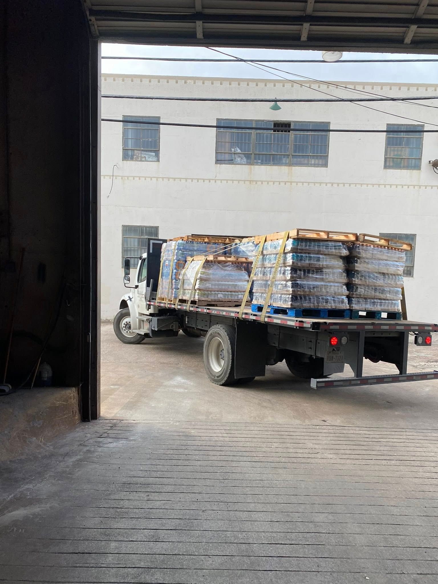 Truck loaded with pallets of goods backed into a loading dock of a white industrial building.