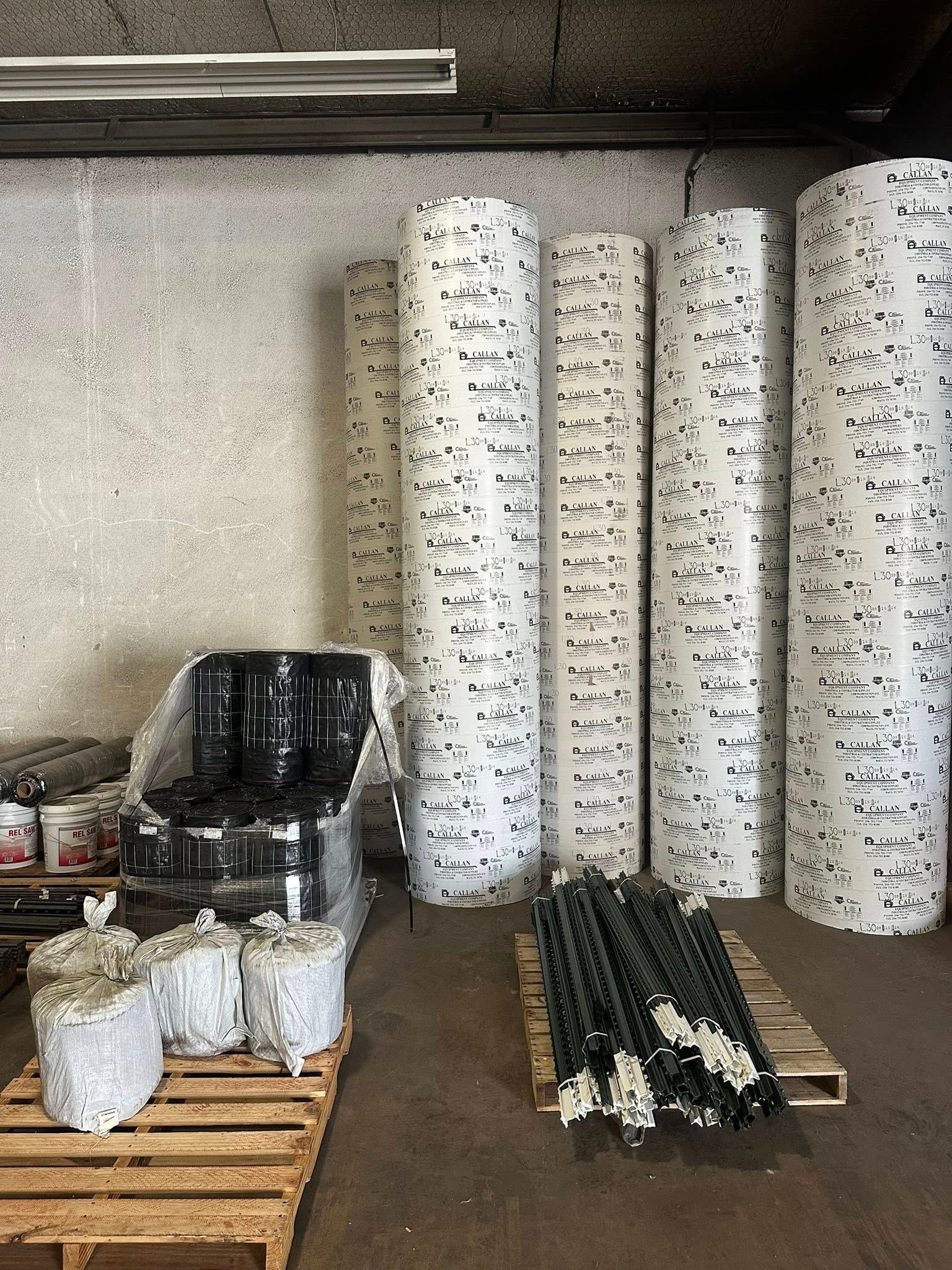 Warehouse with rolls of white material with black speckles, barrels, and pallets.