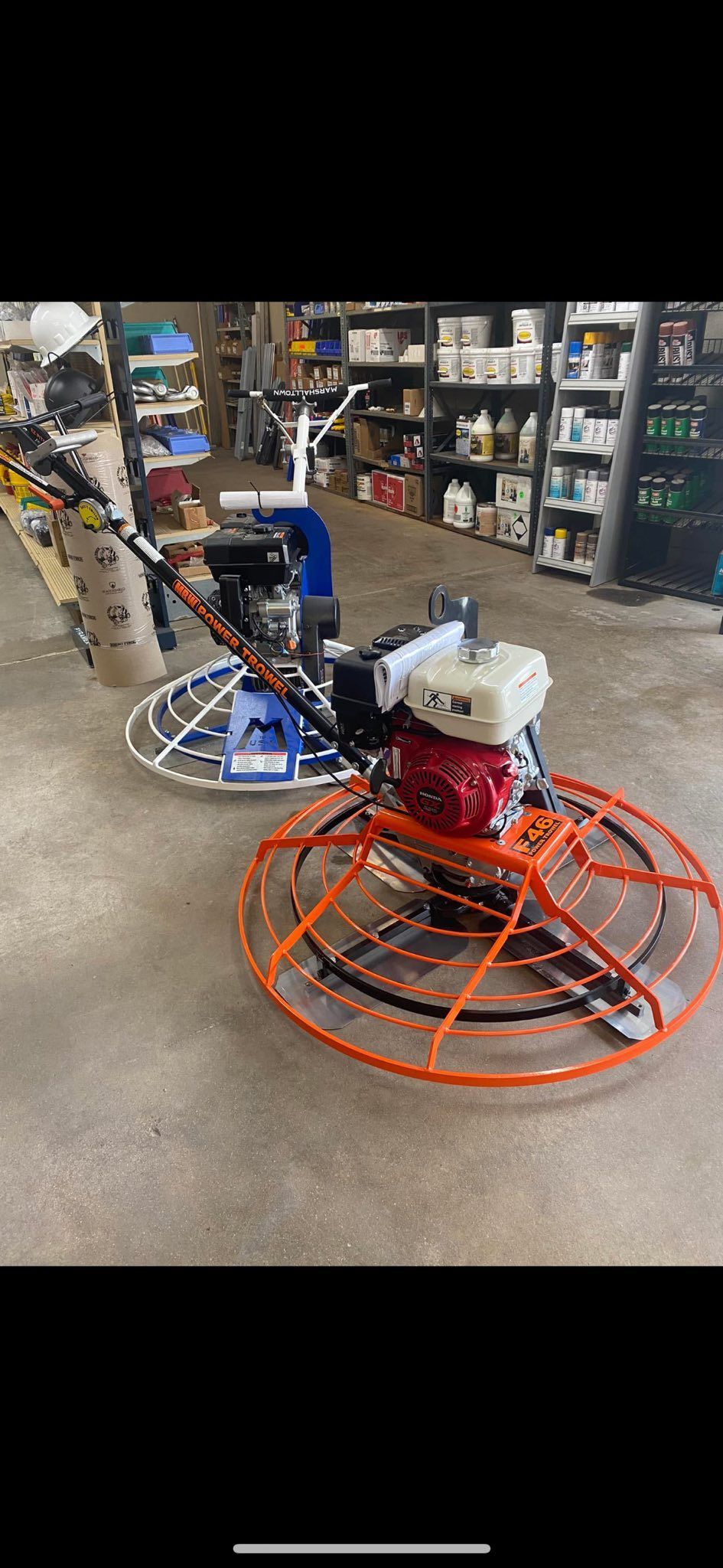 Two concrete finishing machines in a store, one orange and one blue. Shelves of products are in the background.