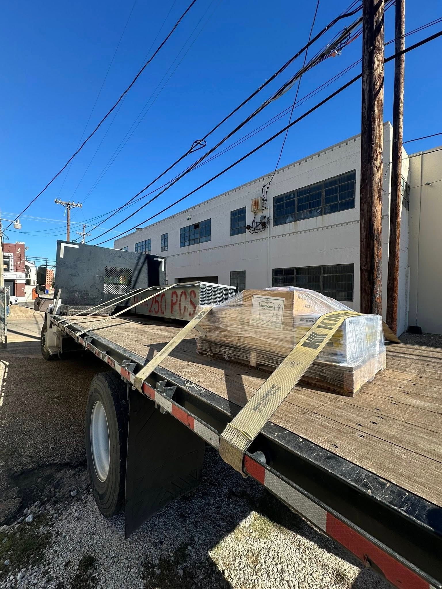 A flatbed truck carrying large, wrapped stone blocks on a sunny day near a white building with power lines overhead.