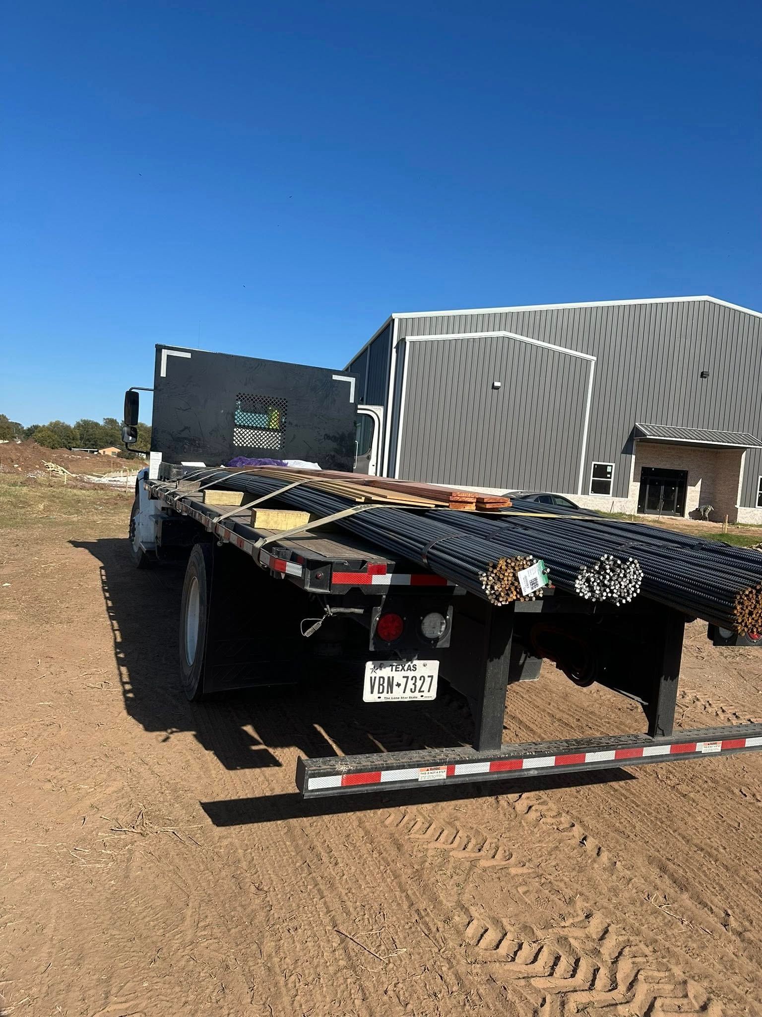 Flatbed truck hauling metal reinforcing bars near a building under a blue sky.