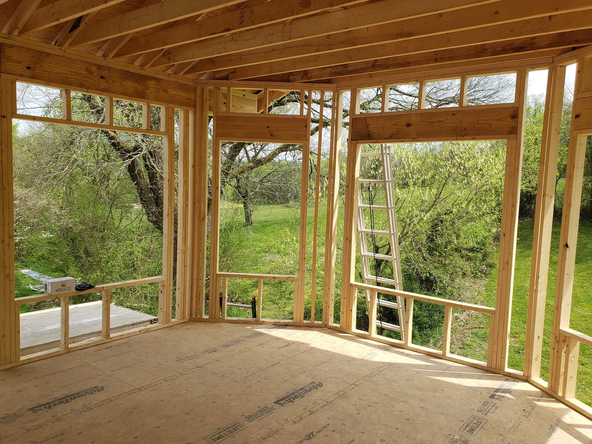 Unfinished wooden porch framing with windows, overlooking green trees and a ladder.