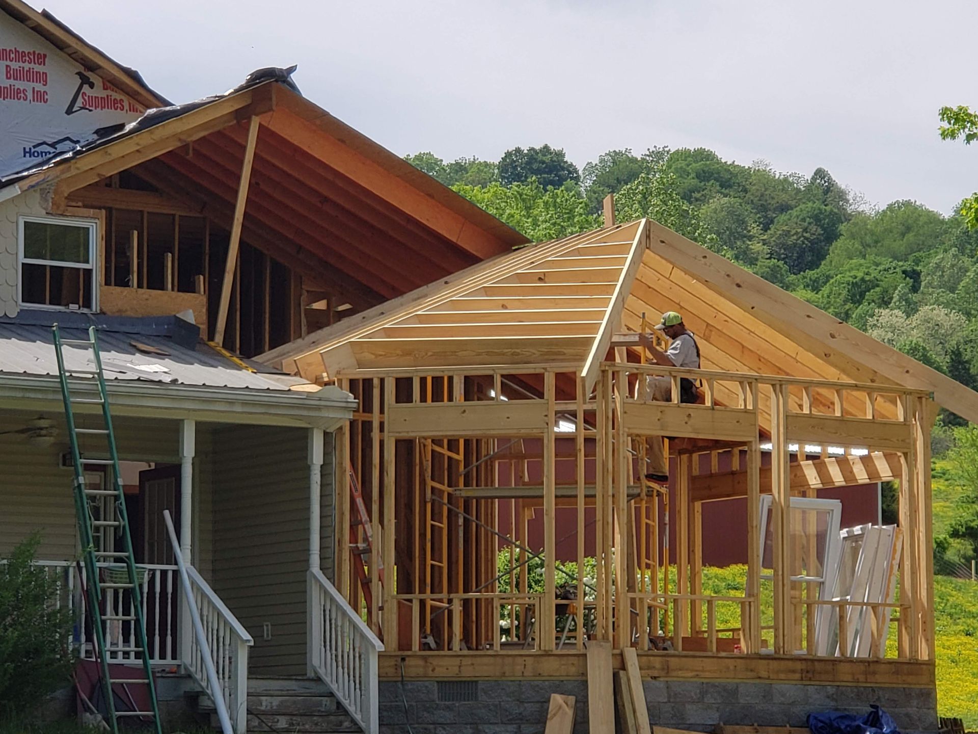 House under construction; a worker on the roof framing. New porch and addition being built in a rural setting.
