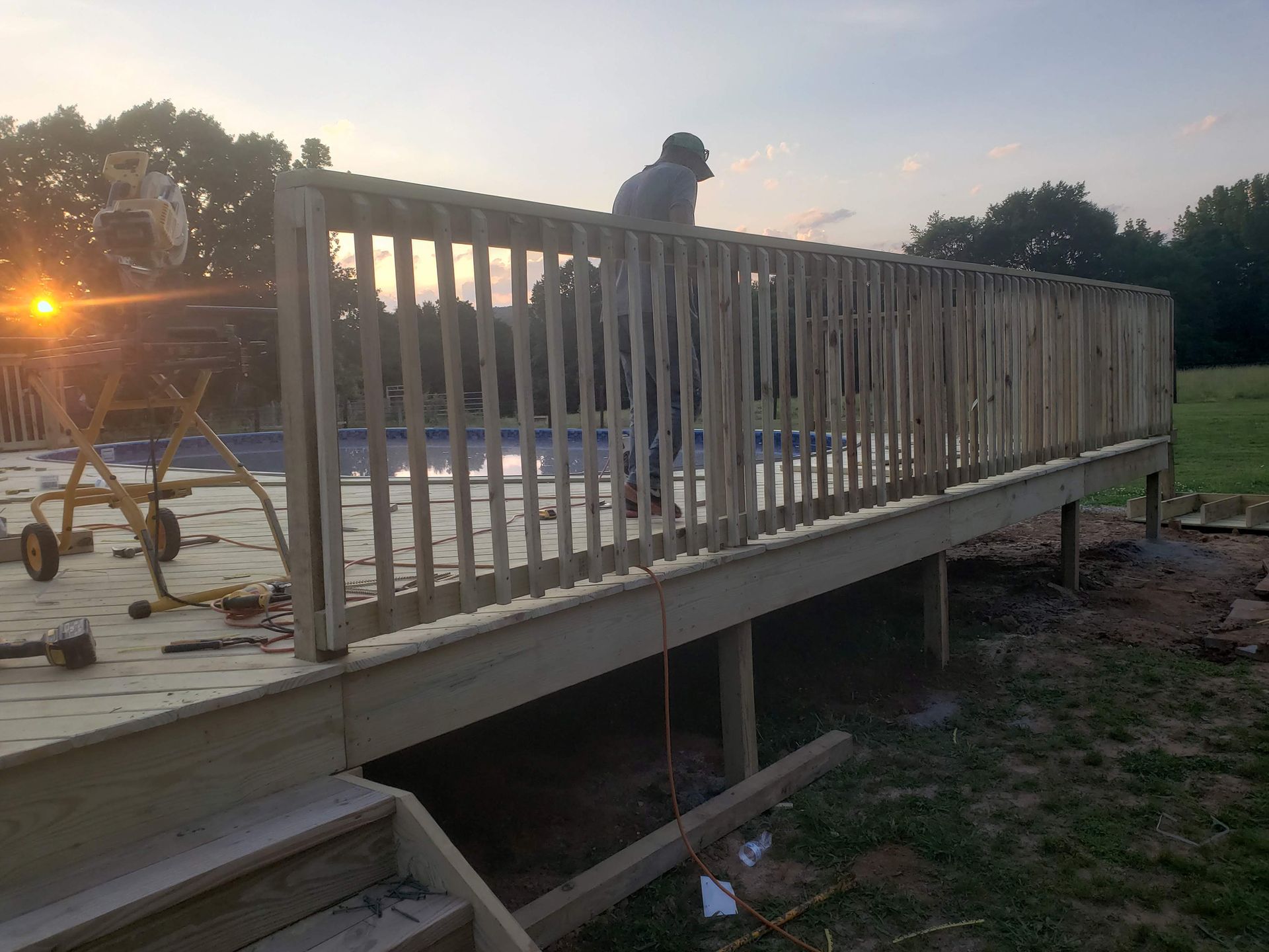 A deck under construction with a person working; in the background is a pool, lawn, and setting sun.