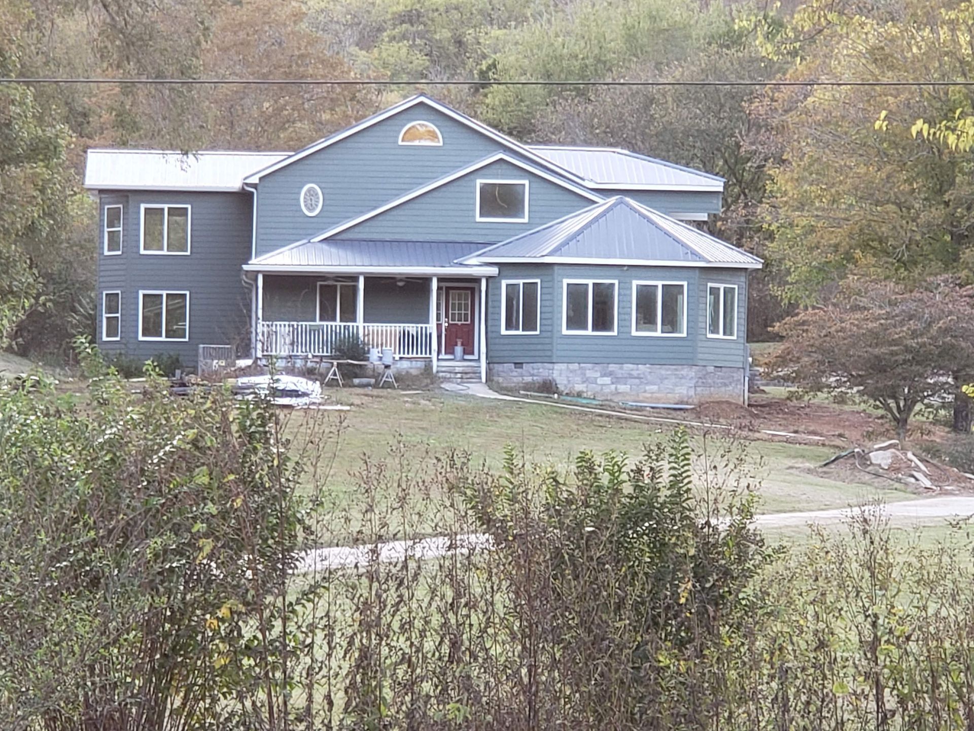 Two-story blue house with metal roof, surrounded by trees and bushes.