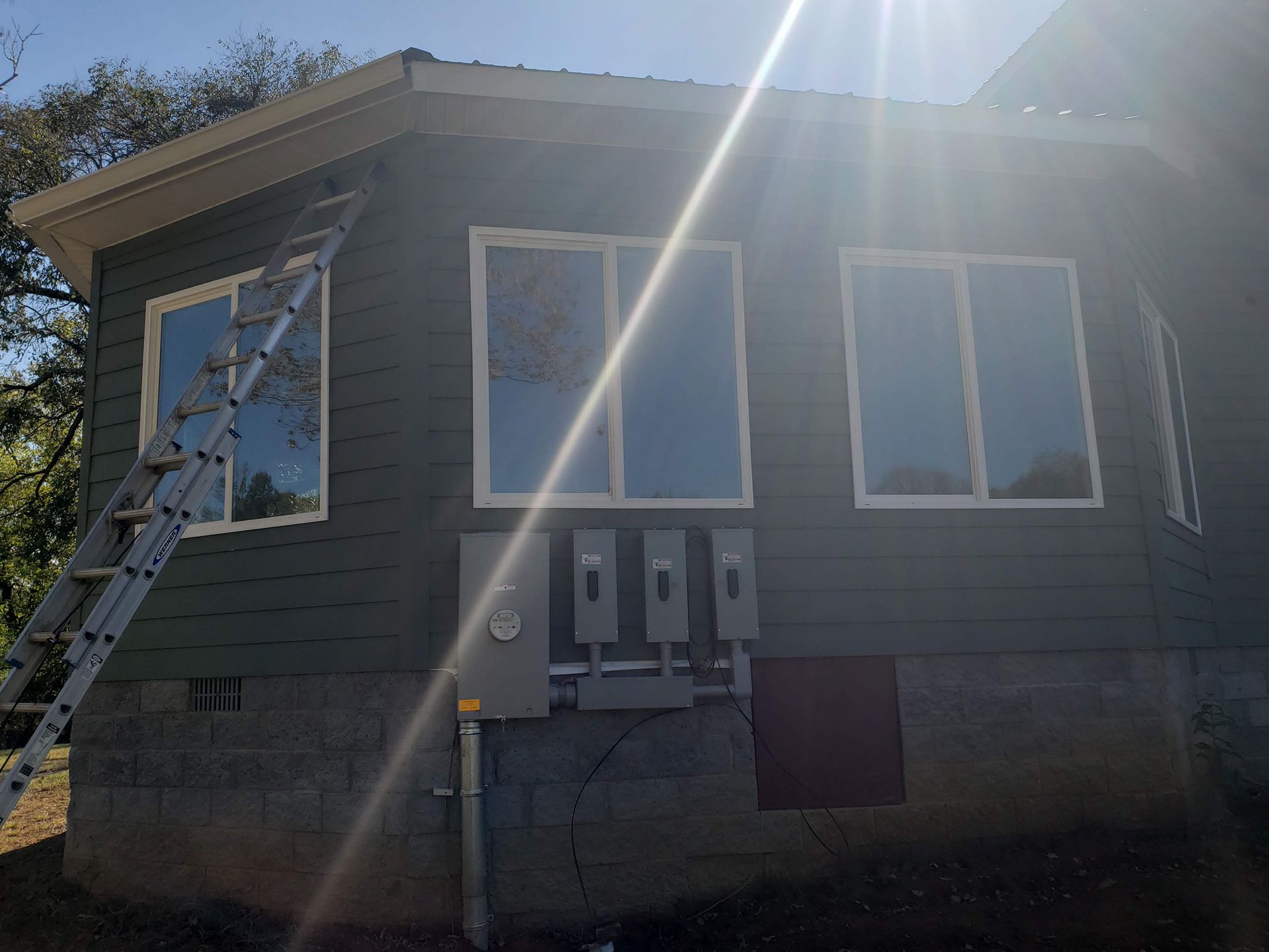 House exterior with green siding, windows, ladder, and electrical box; bright sunlight.