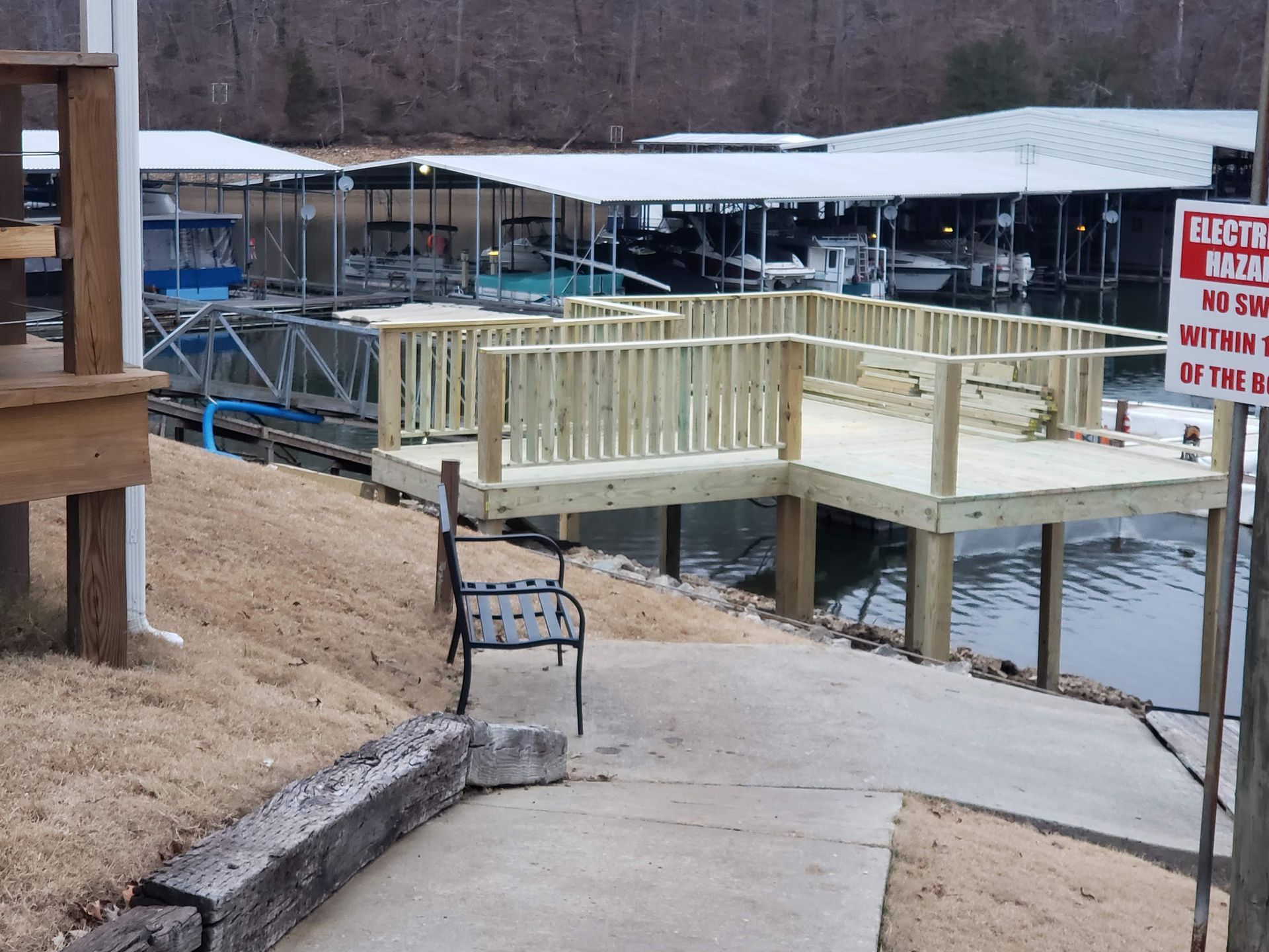 Wooden dock with railings next to a marina, accessible by a concrete path, overlooking water.