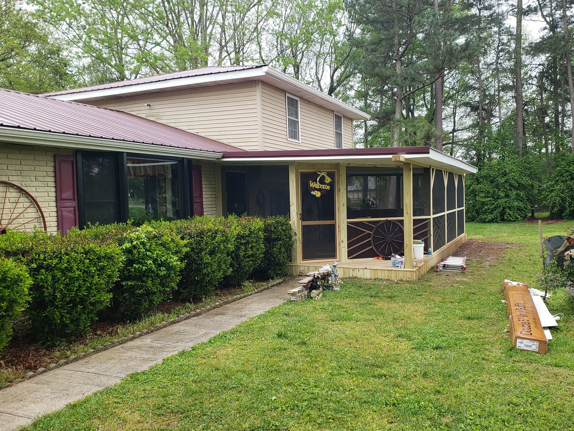 Two-story house with screened-in porch; tan siding, burgundy roof; green lawn, shrubs, and trees in the background.