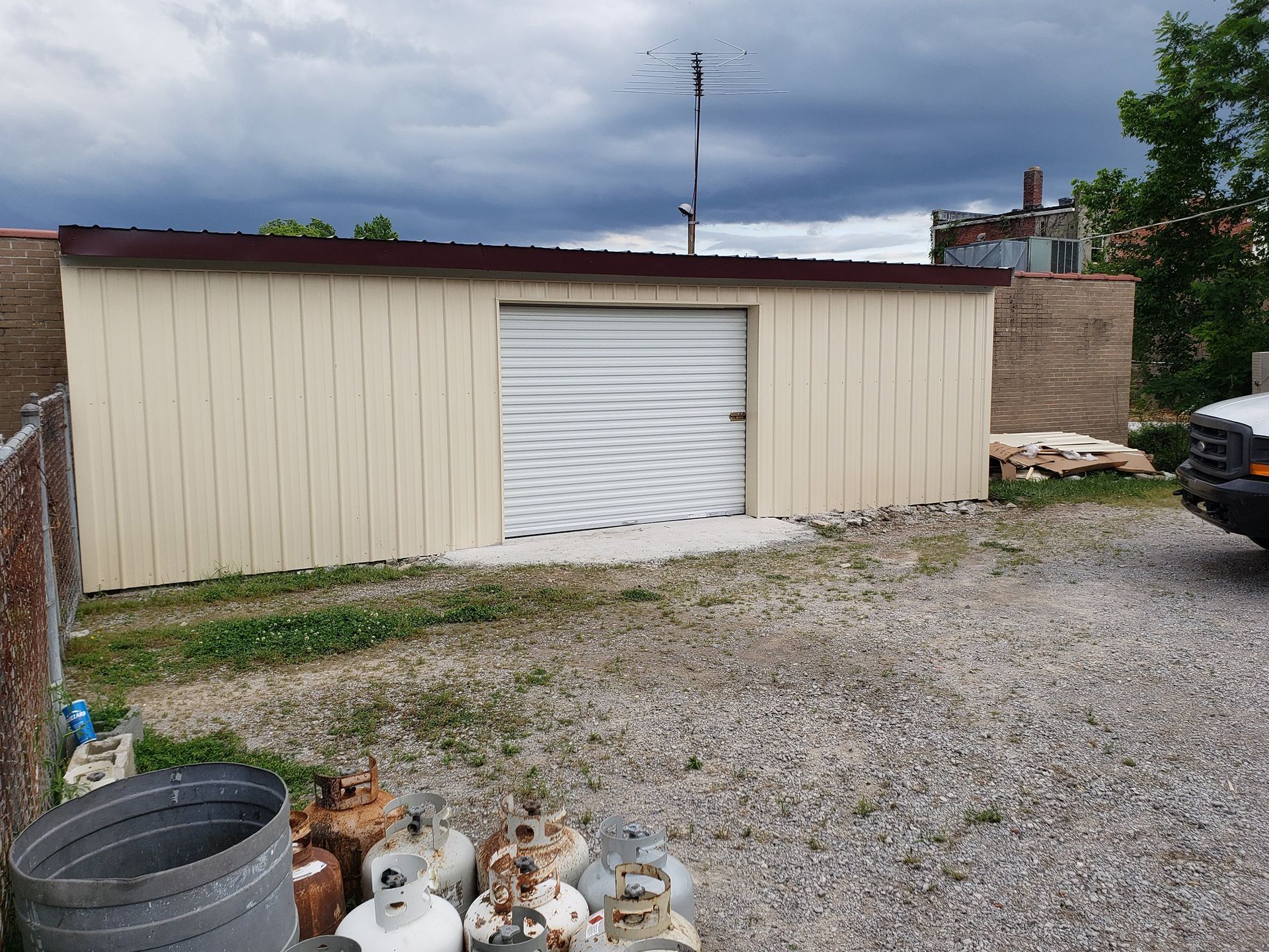 Beige metal shed with roll-up door, gray gravel yard, propane tanks, brick walls, cloudy sky.
