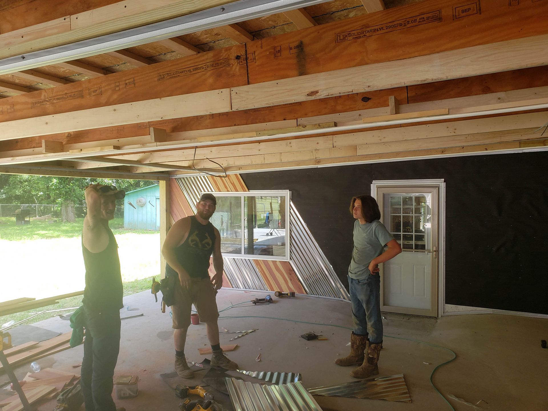 Three people inside a building under construction. One points upwards, others stand and look, corrugated metal on wall.