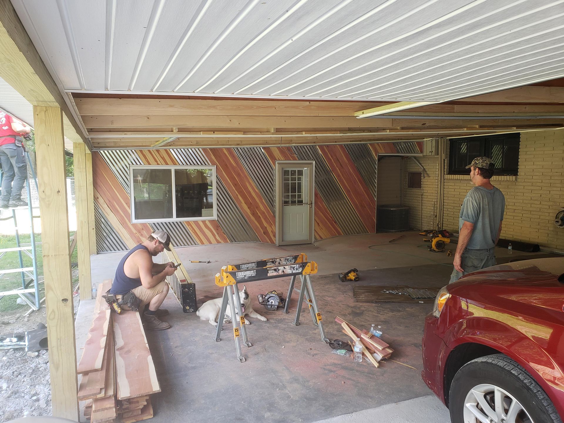 Men working on a building renovation with metal siding, under a carport. One cuts wood, another nearby.
