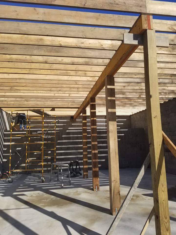 Wooden beams and planks forming a partial roof structure over a concrete surface, with scaffolding visible in the background.