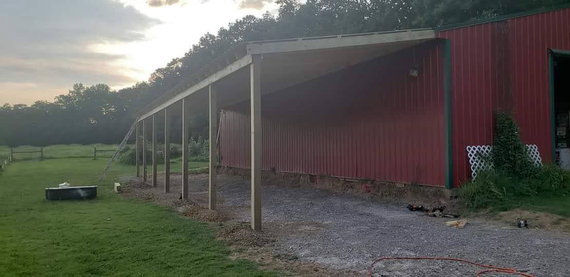 Red barn with a covered walkway supported by wooden posts on a gravel drive, surrounded by green grass and trees.