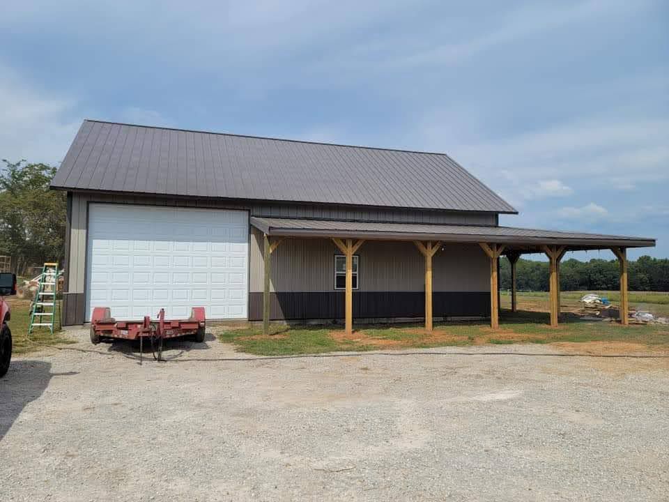 A tan and brown metal building with a lean-to, gravel ground, and a trailer.