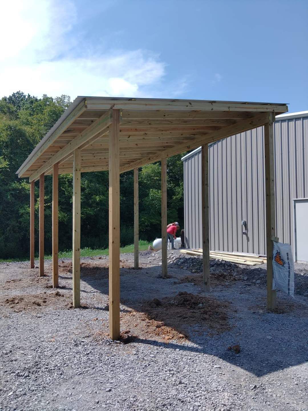 Wooden carport with a sloped roof, erected near a gray metal building on a gravel lot, under a sunny sky.