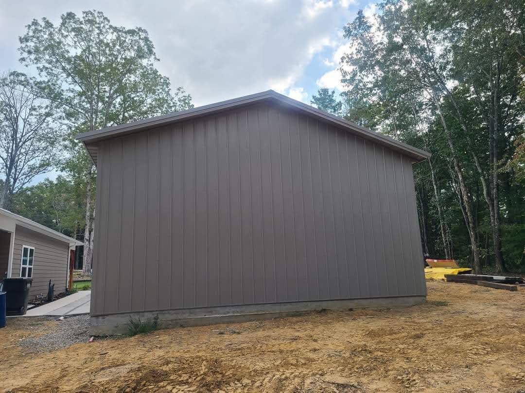 Tan metal-sided shed with a gable roof, set on a lawn, with trees in the background.