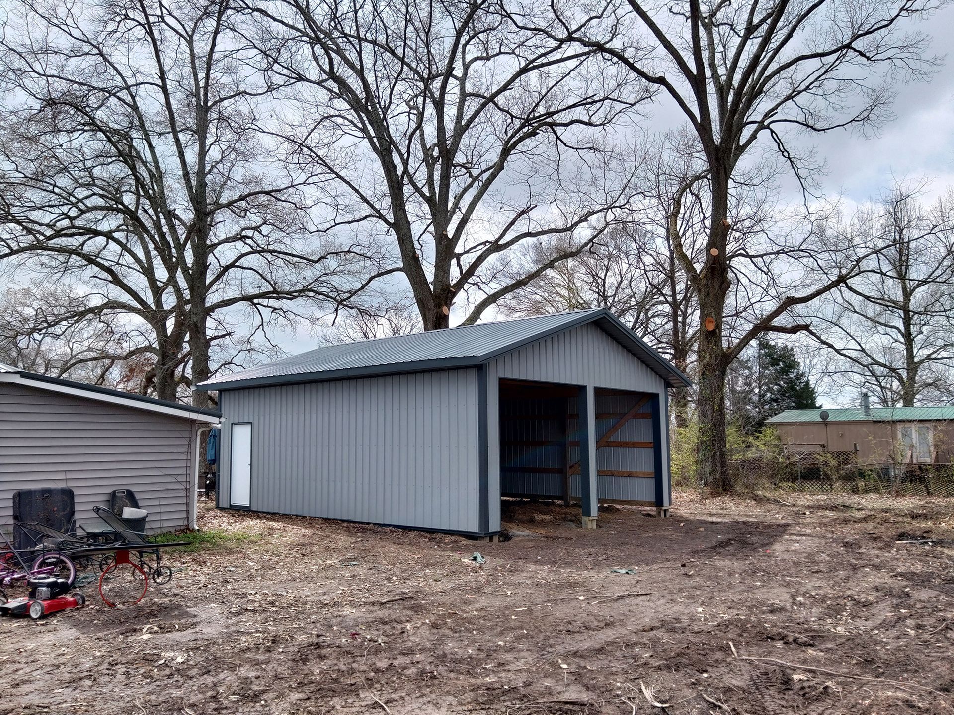 Gray metal garage with two bays and a door, surrounded by leafless trees and a patchy yard.
