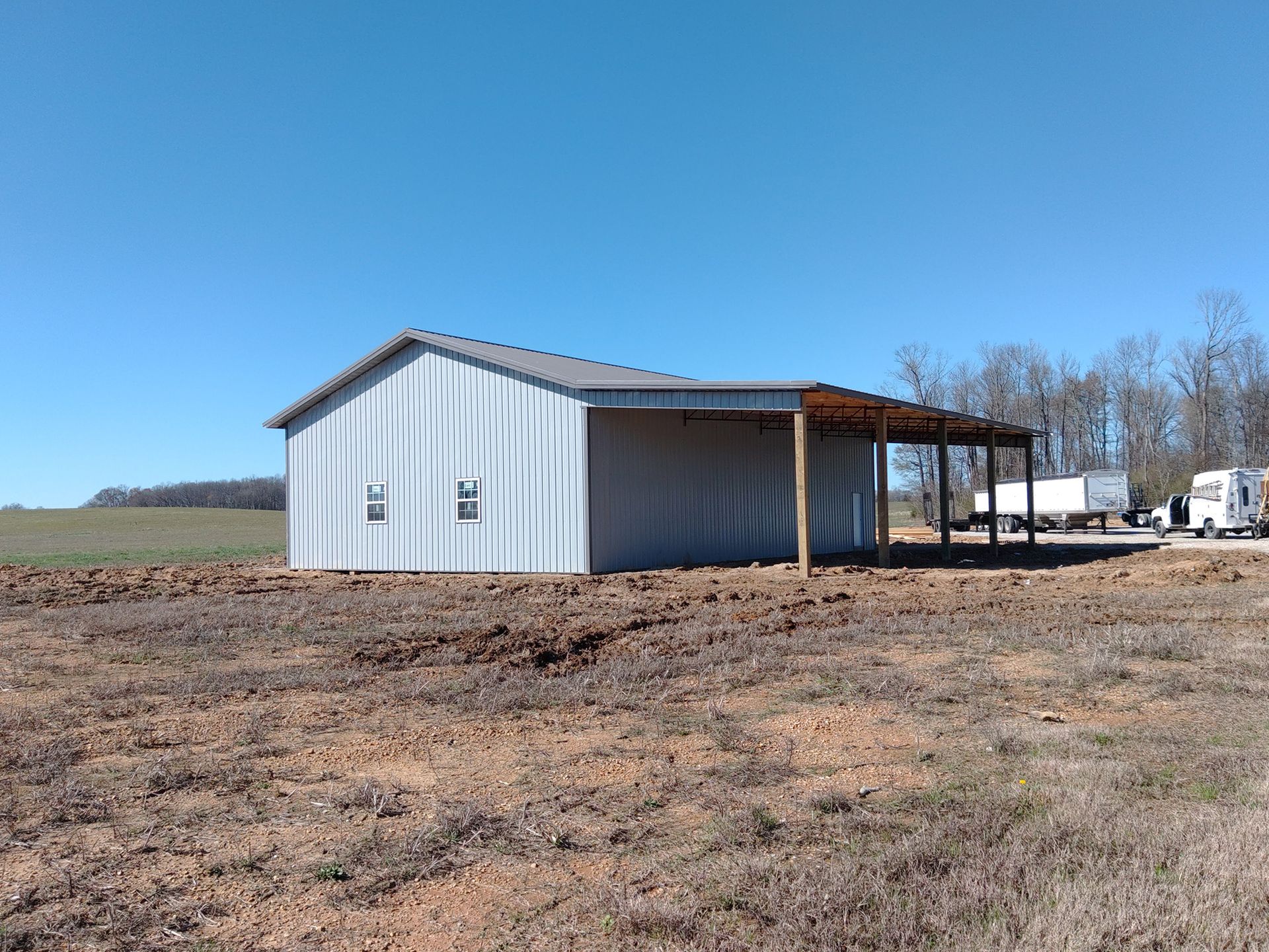 Metal barn with attached open-air structure under a clear blue sky. Located in a field of brown soil.