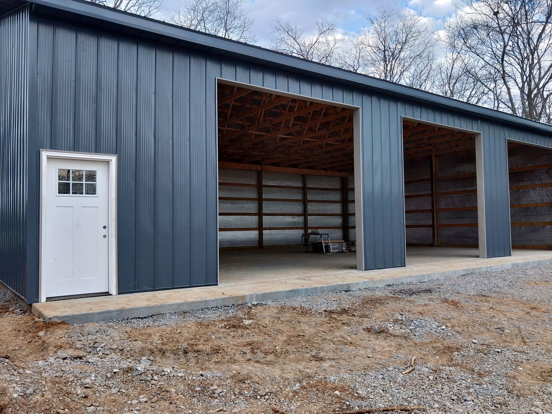 Blue metal building with three bays open to a concrete floor, a white door on the left, and a gravel driveway.