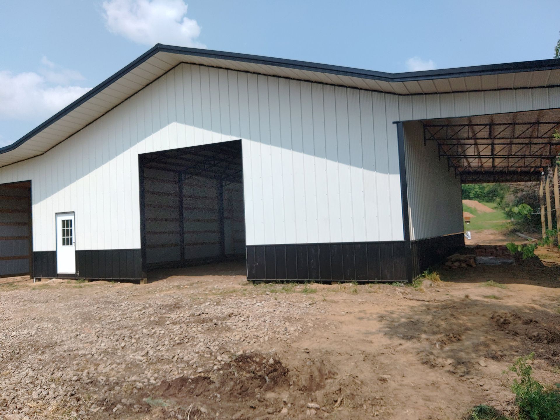 White and black metal agricultural building with open bays and a small door. Gravel and dirt ground.