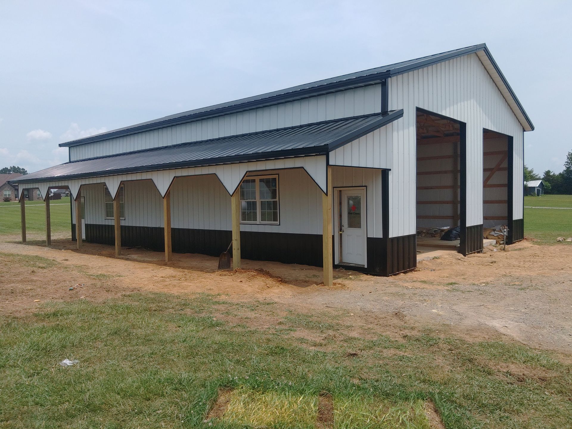 White and dark brown barn with a porch, a black roof, and two open garage bays on a grassy field.