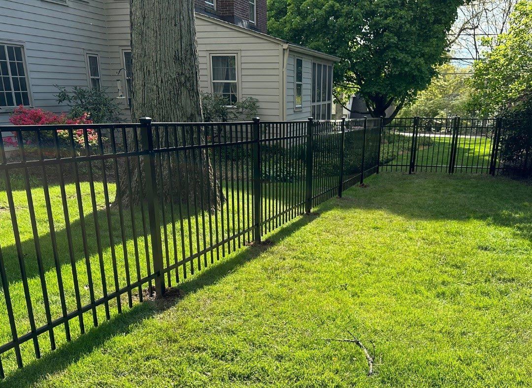 Black metal fence encloses a grassy yard next to a white house with trees in the background.