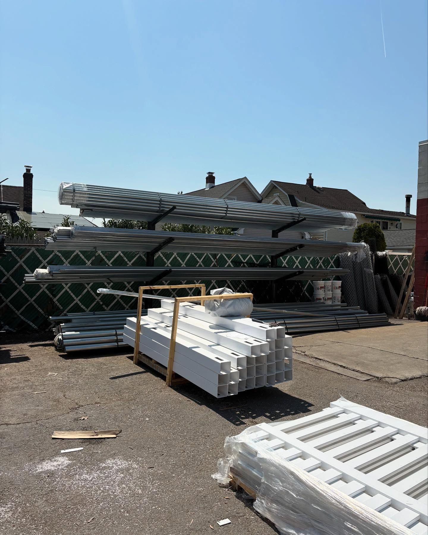 Piles of metal fence parts, including panels and posts, are stacked outside on a sunny day.