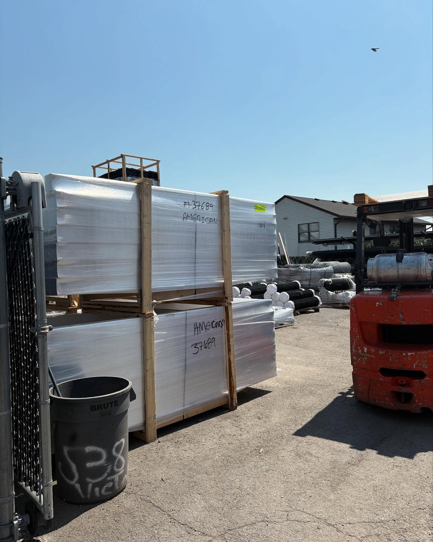 Pallets of wrapped metal, a black bucket, and a forklift in a sunny outdoor area.