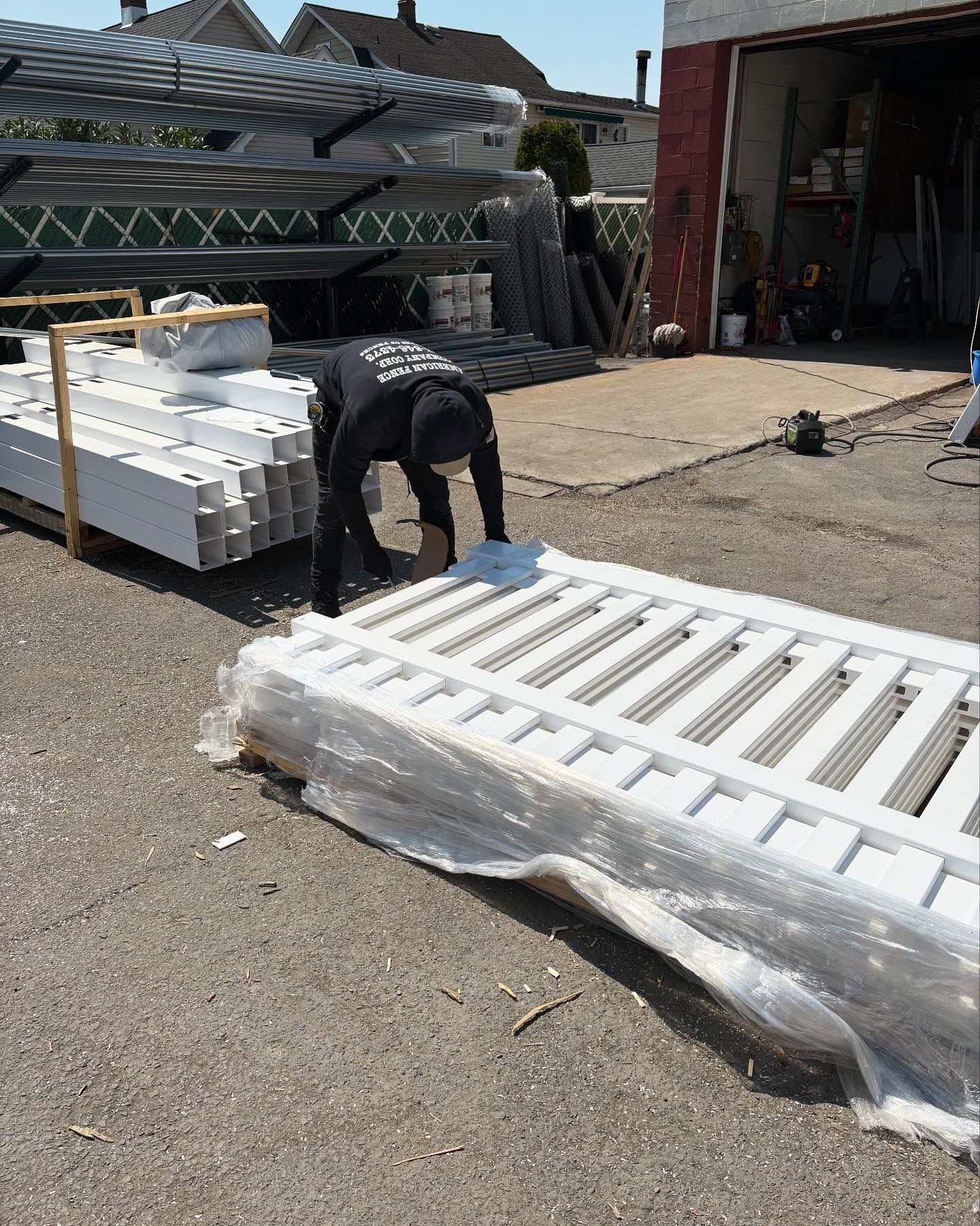 Person in black unloading white fencing. Outdoors, pallets, and garage visible.