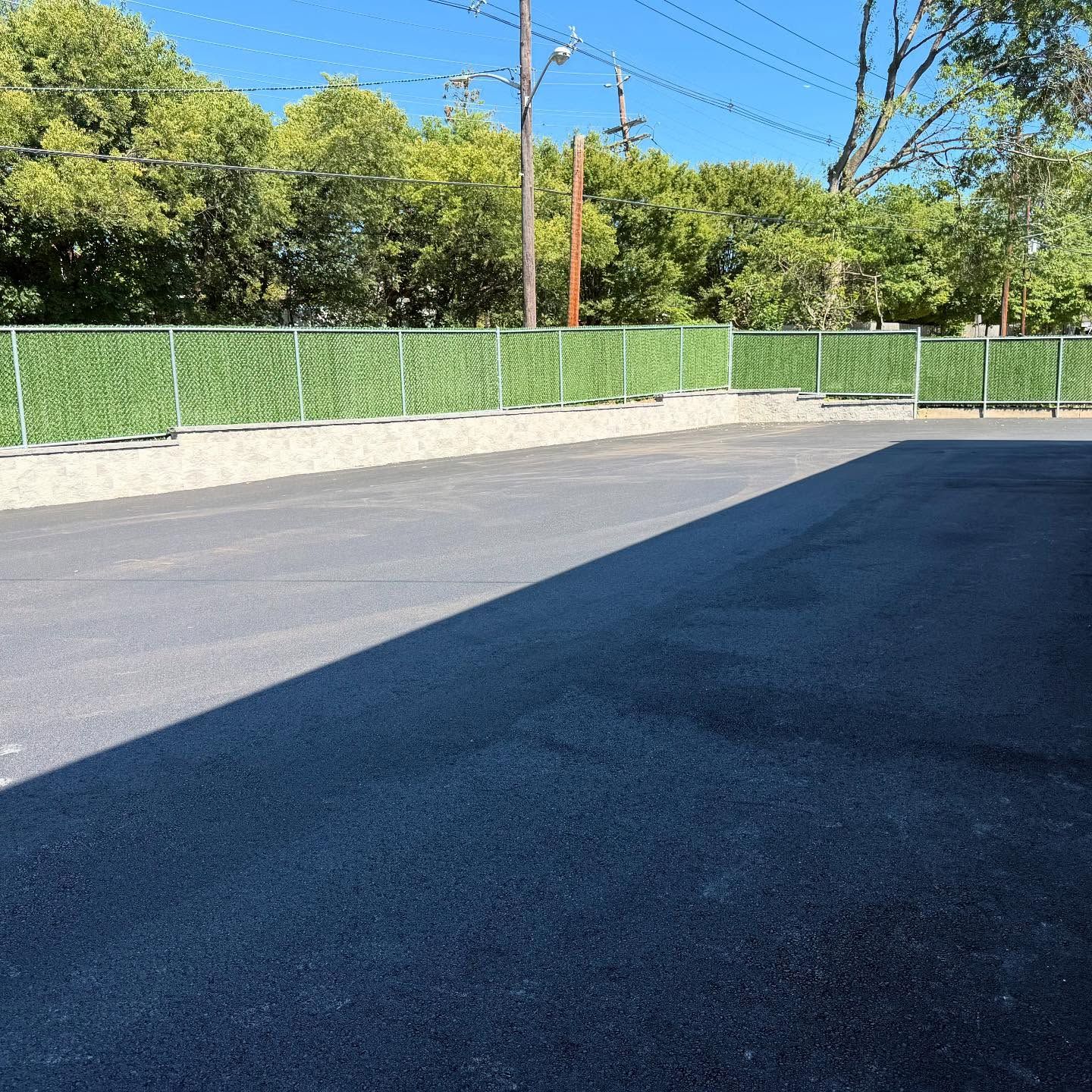 Asphalt parking lot with a chain-link fence covered in green artificial foliage and a row of trees.