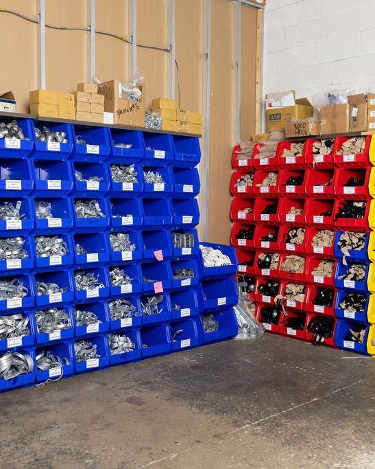 Blue, red, and yellow storage bins filled with parts are stacked in a warehouse. Cardboard boxes sit on top.