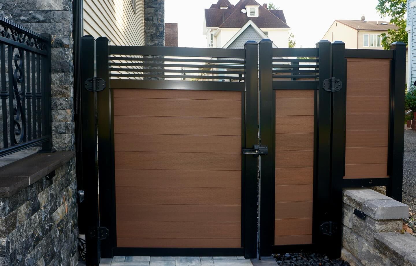 Brown and black wooden gate with horizontal slats, leading to a house, set in a brick-paved area.