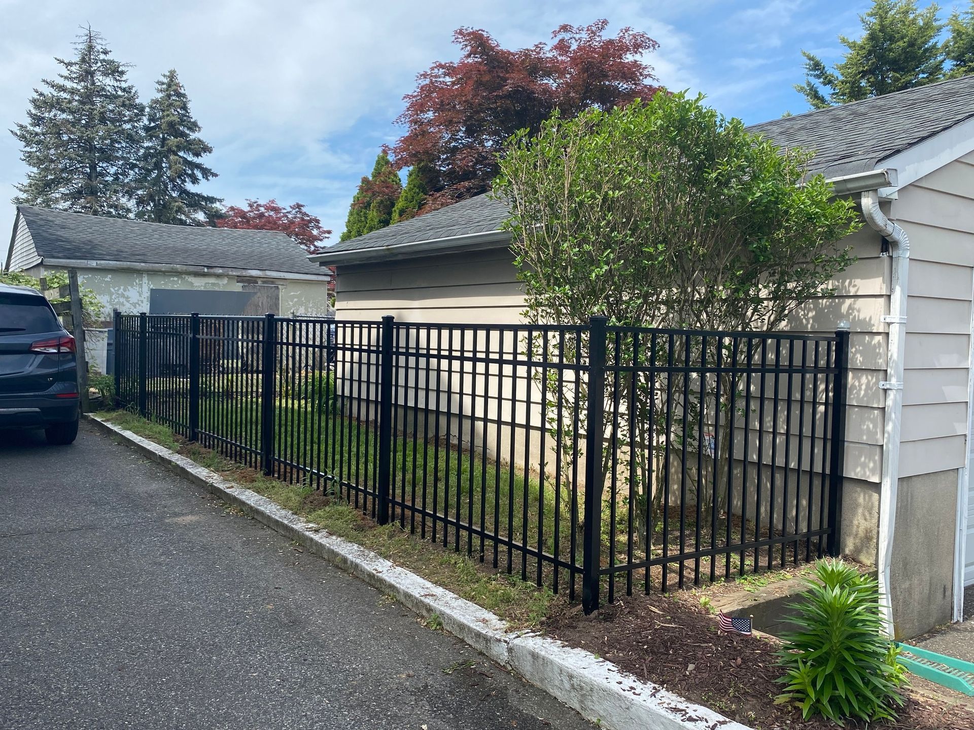 Black metal fence along a driveway next to a garage, with green foliage and a blue sky.