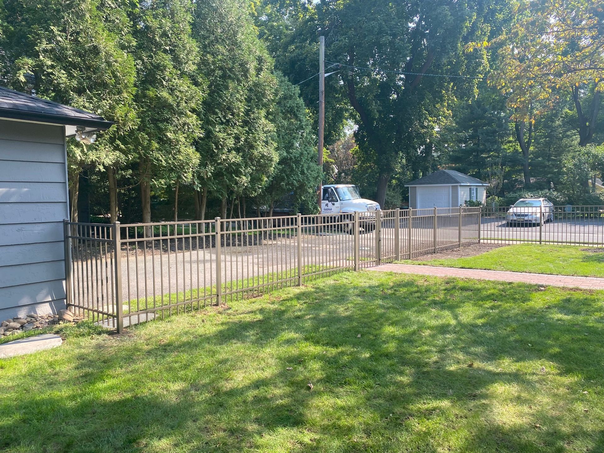 A metal fence surrounds a grassy yard, with a small building on the left and cars parked beyond.