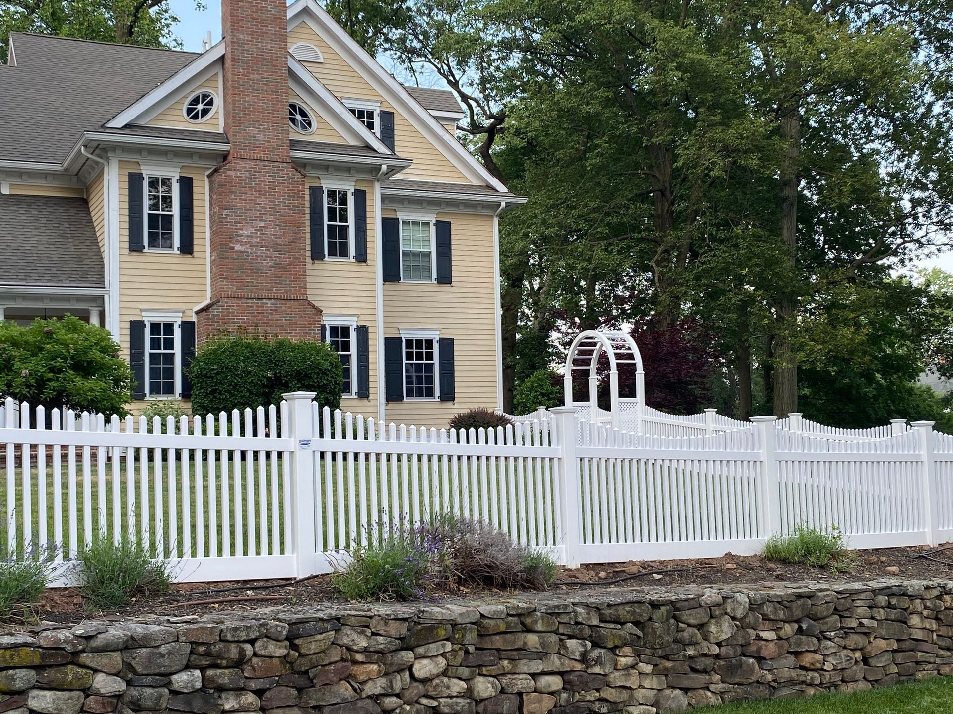 White picket fence in front of a yellow house with black shutters and a stone retaining wall.