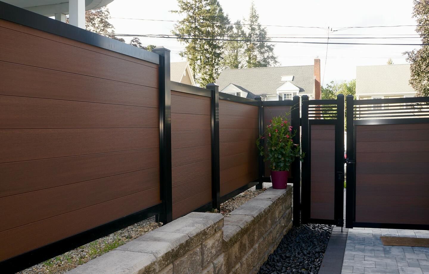 Brown and black composite fence with gate in front of a house, gravel and retaining wall in the foreground.