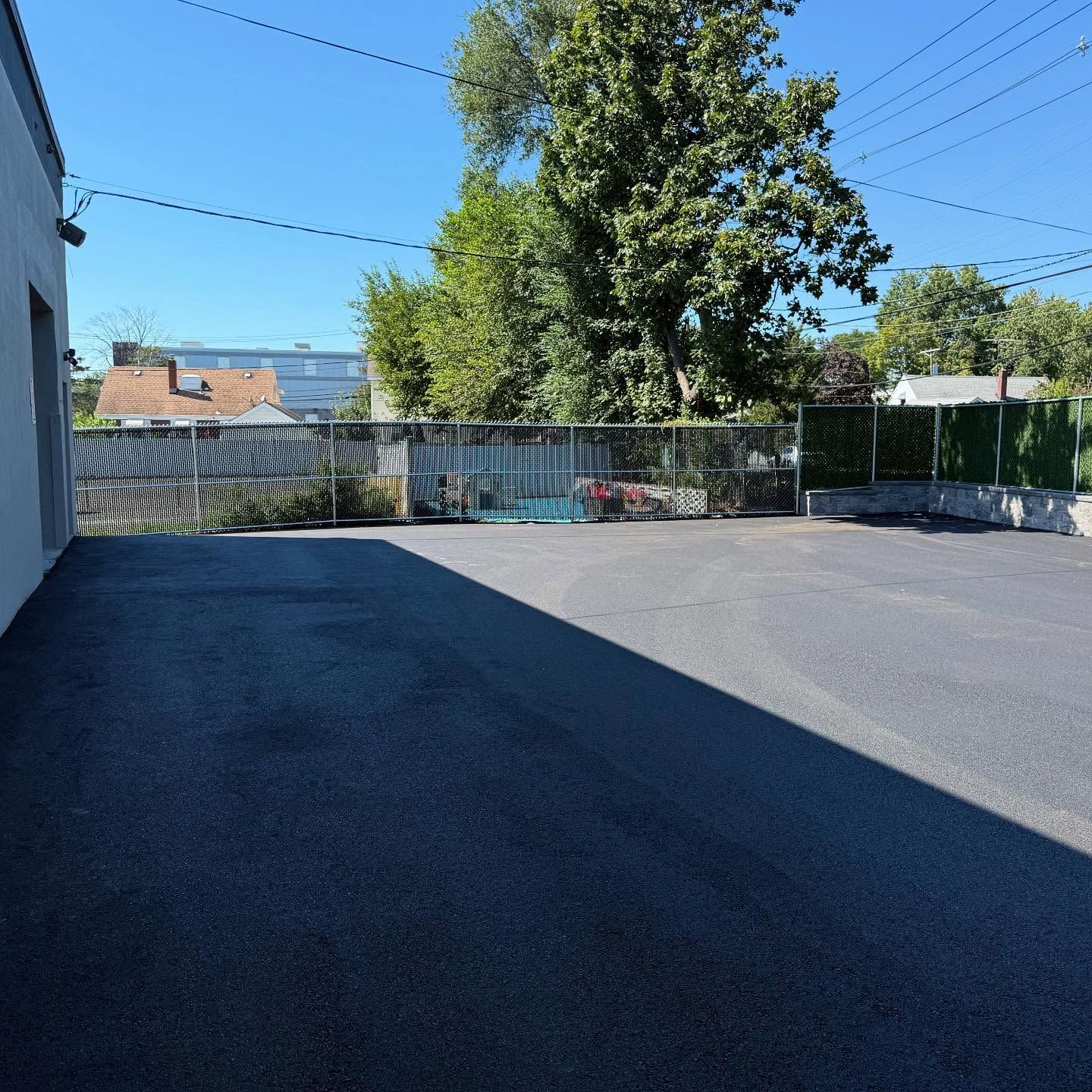 Empty asphalt lot with a gray building on the left, fence, and trees under a clear blue sky.