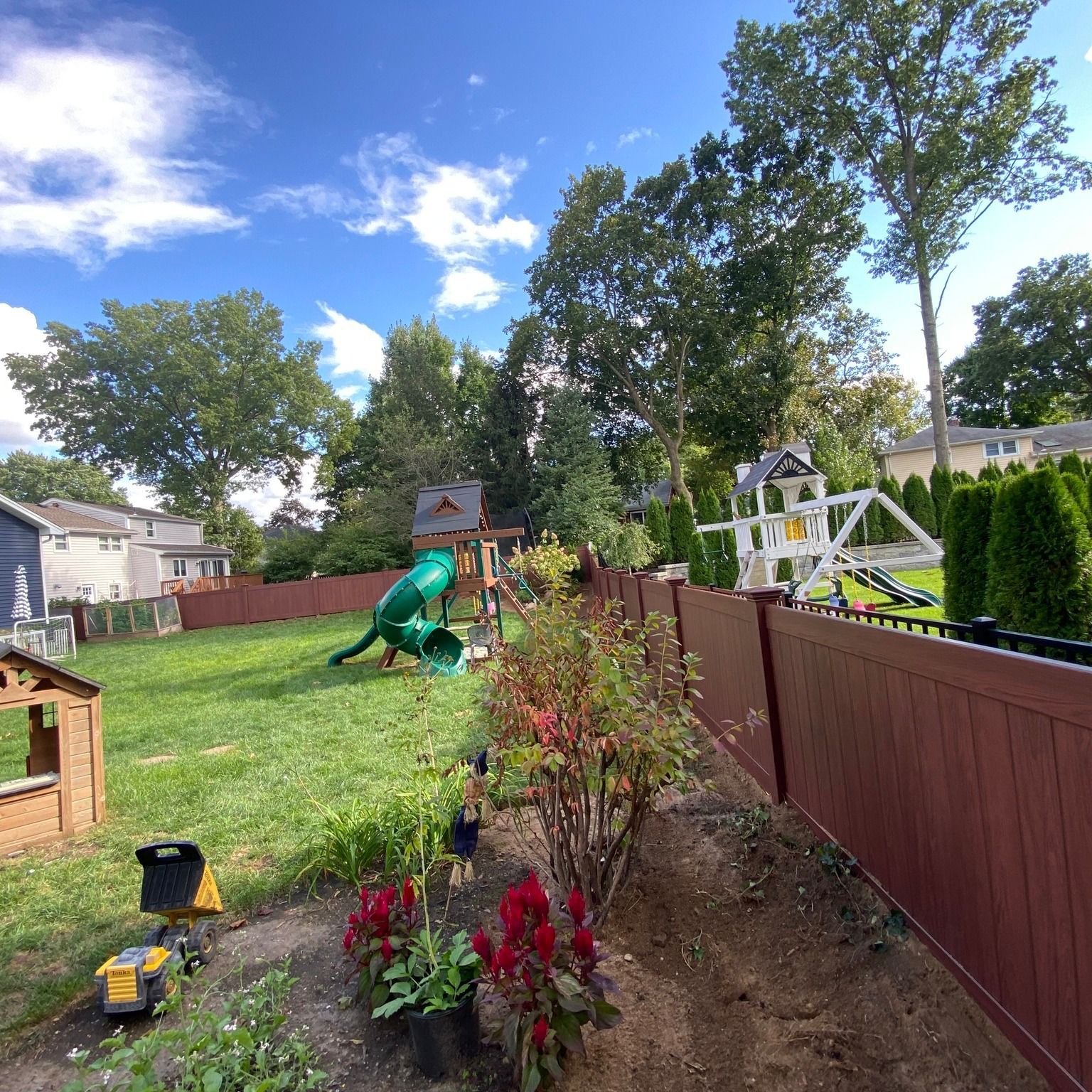 Backyard with red fence, grass, playground, swing set, trees, and blue sky.
