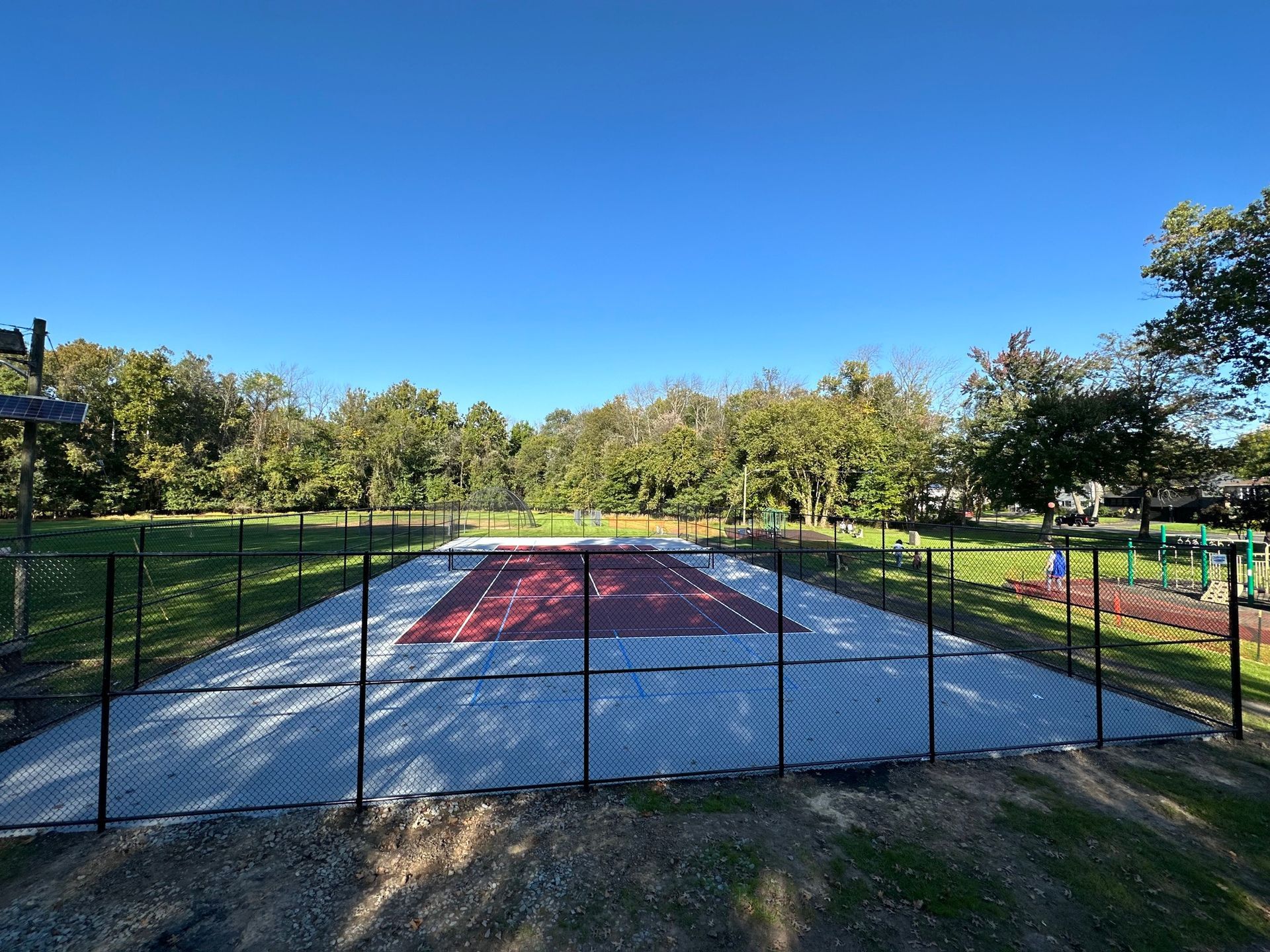 Fenced pickleball court; red and gray surface; green trees and grass in the background under a blue sky.