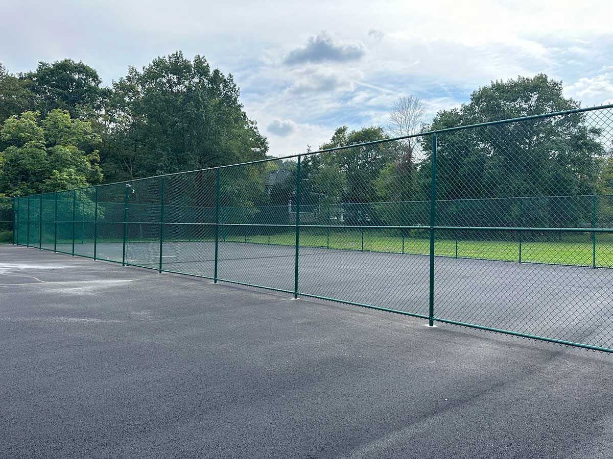 Green chain-link fence surrounding a gray asphalt surface, with trees in the background under a cloudy sky.